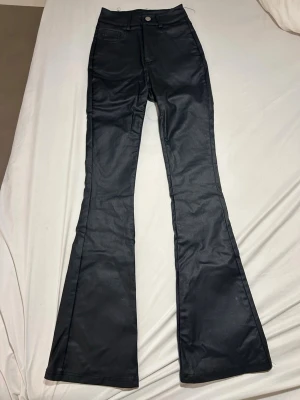 Svarta utsvängda jeans - Snygga svarta jeans med hög midja och utsvängda ben. Jeansen har klassiska fickor och stängs med knapp och dragkedja. Materialet har en lätt glansig finish som ger en cool look. Perfekta för dig som gillar trendiga och bekväma byxor.