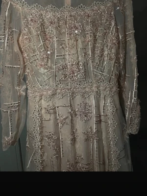 Lång ljusrosa spetsklänning med broderi - Magisk långklänning i ljusrosa med transparenta partier och vackra broderade blommor samt paljettdetaljer. Klänningen har långa ärmar i tunn mesh och markerad midja. Perfekt för speciella tillfällen och ger en riktigt sagolik känsla.