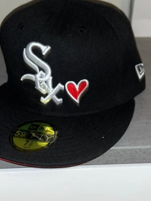 Svart New Era White Sox keps med hjärta - Säljer en svart keps från New Era, modell 59FIFTY, med White Sox-logga och broderat rött hjärta framtill. Kepsen har platt skärm och broderad detalj med hjärta även på sidan. Perfekt för dig som vill sticka ut med stilren streetwear.