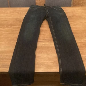Mörkblå jeans från DAKING - Snygga mörkblå jeans från DAKING med klassisk femficksdesign och kontrastsömmar. Jeansen har raka ben och silverfärgade detaljer, inklusive en metallbokstav vid fickan. Perfekta för dig som gillar stilrena och tidlösa jeans.
