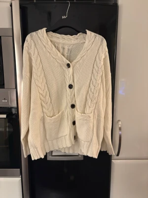 Vit kabelstickad kofta med fickor - Mysig vit kabelstickad kofta med stora svarta knappar framtill och två praktiska fickor. Koftan har ribbade muddar och ett avslappnat snitt. Perfekt för lager på lager och ger en chill vibe till din outfit.