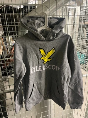 Grå hoodie från Lyle & Scott - Snygg grå hoodie från Lyle & Scott med stor gul fågel-logga och vit text på bröstet. Klassisk känguruficka framtill och huva. Perfekt för dig som gillar streetwear och vill ha en stilren look.