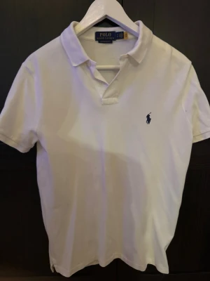Vit pikétröja från Polo Ralph Lauren - Klassisk vit pikétröja från Polo Ralph Lauren med blå broderad logga på bröstet. Tröjan har krage, korta ärmar och en slim passform. Perfekt till jeans eller shorts för en clean och stilren look. (Pris kan diskuteras)