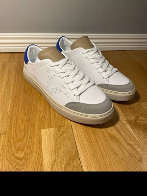 Vitt sneakers från Versace 19.69 - Snygga vita sneakers från Versace 19.69 med blå detalj på hälen, beige plös och grå förstärkning vid tån. Skorna har perforerad sidopanel och klassisk låg profil. Tillverkade i läder med gummisula och vita skosnören.