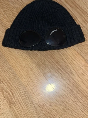 Svart goggle-mössa från C.P. Company - Säljer en svart ribbstickad mössa från C.P. Company med de ikoniska goggle-linserna framtill. Mössan är i mjukt och stretchigt material och passar perfekt för dig som vill ha en streetwear-look med attityd.