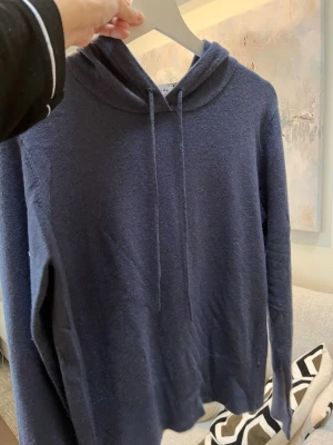 Mörkblå hoodie kashmir  - Mysig mörkblå hoodie med huva och dragsko. Tillverkad i ett mjukt, stickat material som känns skönt mot huden. Perfekt för en avslappnad och stilren look. Långärmad och enkel design utan tryck eller logga.