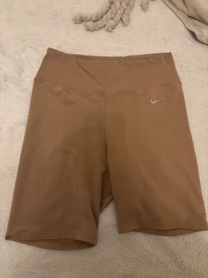 Bruna cykelshorts från Nike - Snygga bruna cykelshorts från Nike med hög midja och diskret logga framtill. Perfekta för träning eller en sportig look. Stretchigt material som sitter skönt och följsamt.