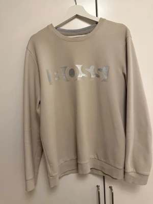Beige sweatshirt från BOSS - Snygg beige sweatshirt från BOSS med silverfärgad logotyp på bröstet. Tröjan har rund halsringning, ribbade muddar och är tillverkad i mjukt material som känns skönt mot huden. Perfekt för en avslappnad och trendig look. Knappt använt väldigt bra skick