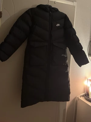 Svart lång dunjacka från Nike - Säljer en svart lång dunjacka från Nike med huva och dragkedja framtill. Jackan har en oversized look, två fickor med lock och diskret Nike-logga på bröstet. Perfekt för kalla vinterdagar och riktigt varm tack vare dunfyllningen.