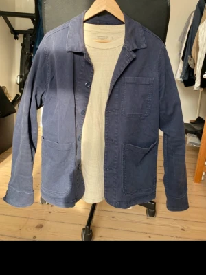 Marinblå overshirt från Universal Works - Säljer en marinblå overshirt från Universal Works i mjuk bomull. Jackan har tre stora utanpåfickor, knäppning framtill och klassisk krage. Perfekt lager-på-lager-plagg med avslappnad passform och stilren look.