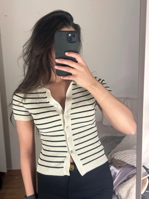 Randig ribbad kortärmad topp - Snygg ribbad topp med korta ärmar och smal passform. Toppen är vit med svarta horisontella ränder, har en klassisk krage och knappar framtill. Perfekt till jeans eller kjol för en trendig look. Petit S 
