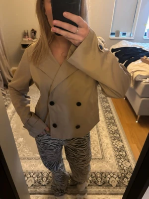 Kort beige trenchcoat från Vila - Säljer min trenchcoat från vila då jag inte använt den så mycket och den kommer ej komma till användning! Den är jätte fin till vår/höst men har en liten fläck som ej syns när man knäpper jackan.  Köparen kan försöka ta bort den( jag har ej försökt noga utan bara tvättat den)
