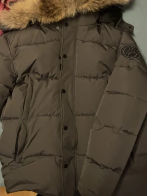 Brun dunjacka från Canada Goose - Snygg brun dunjacka från Canada Goose med stor huva och fuskpäls. Jackan har en klassisk puffer-modell, dragkedja och knappar framtill samt två stora fickor. Märkets logga syns på soparmen. Perfekt för kalla dagar och riktigt varm.