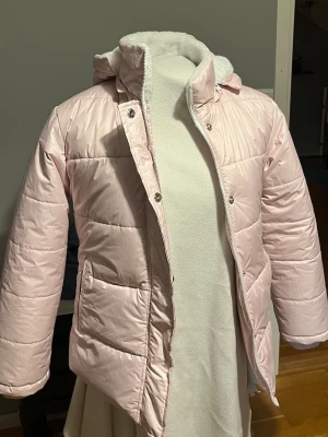 Ljusrosa pufferjacka med huva - Säljer en ljusrosa pufferjacka med huva och dragkedja. Jackan har quiltad design, två sidofickor och mjukt vitt foder på insidan av kragen. Perfekt för kalla dagar och riktigt mysig att bära. Fick den i julklapp men aldrig använd då den var för liten för mig💕