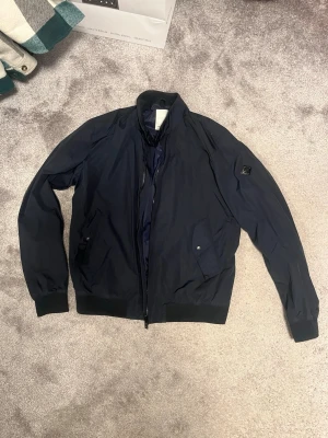 Mörkblå bomberjacka från J.lindeberg - Snygg mörkblå bomberjacka från J.LINDEBERG med ribbade muddar, dragkedja framtill och två fickor med tryckknappar. Jackan har en klassisk patch på ärmen och är tillverkad i ett lätt, vindtätt material.  Storlek L| enda flaw är en fickknapp är borta 