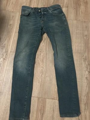 Blå slim fit jeans  - Snygga blå slim fit jeans från Cotton Smith med klassisk femficksdesign och knappgylf. Jeansen har en lätt tvättad look med slitningar på låren och bakfickor med diskreta sömmar. Perfekta för en avslappnad och trendig stil.