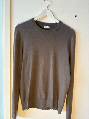 Brun stickad tröja från Filippa K - En stilren grön/grå stickad tröja från Filippa K med rund halsringning och långa ärmar. Tröjan har en enkel, klassisk design med ribbade muddar vid ärmslut och nederkant. Perfekt för lager på lager och passar till det mesta.