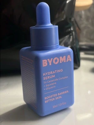 BYOMA Hydrating Serum blå flaska - BYOMA Hydrating Serum i en blå, fyrkantig plastflaska med pipett. Innehåller Tri-Ceramide Complex, squalane och glycerin för att boosta hudbarriären. Flaskan rymmer 30 ml och har rosa text på framsidan. Perfekt för dig som vill ha återfuktad och fräsch hud. Testad en gång på handen. Orginal pris: 199kr mitt pris: 170