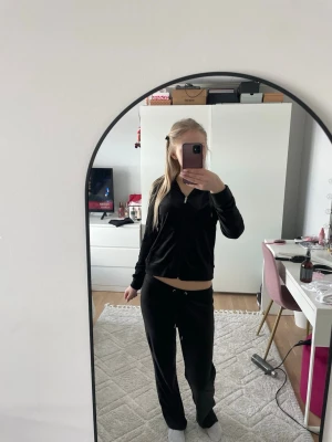 Svart velour loungewear-set - Mysigt svart loungewear-set i velour med zip-tröja och matchande byxor med dragsko i midjan. Perfekt för chill hemma och har en klassisk, enkel design. Tröjan har långa ärmar och byxorna är raka och mjuka.