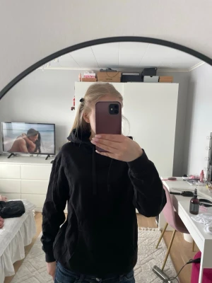 Svart hoodie med huva - En klassisk svart hoodie med huva och dragsnören. Tröjan har en stor magficka framtill och ribbade muddar vid ärmslut och nederkant. Perfekt för en avslappnad och cool look. Materialet ser ut att vara mjukt och bekvämt, troligen bomullsblandning.