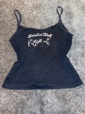 Svart Howlin' Wolf linnetopp - Cool svart linnetopp med smala axelband och tryck med texten 'Howlin' Wolf' framtill. Toppen har en figurnära passform och är perfekt för en avslappnad och trendig stil.