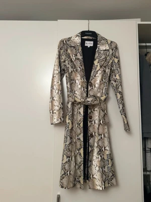 Ormmönstrad kappa från NA-KD - Snygg lång kappa från NA-KD med ormmönster i beige, brunt och svart. Kappan har klassisk krage, knäppning framtill och tillhörande bälte i midjan. 