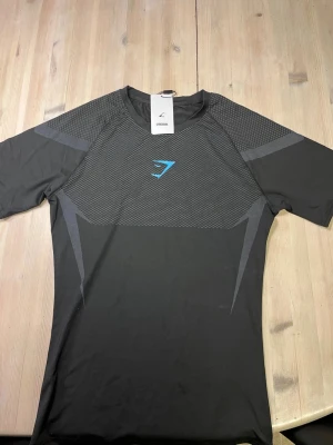 Gymshark onyx svart Compression shirt med lappen kvar - Snygg svart t-shirt från Gymshark med gråa detaljer och ett blått logotryck på bröstet. Tröjan har korta ärmar och är tillverkad i ett stretchigt syntetmaterial som passar perfekt för träning. Modern design med mönstrade partier på både fram- och baksida. / svår att få tag på. Lappen sitter fortfarande på!