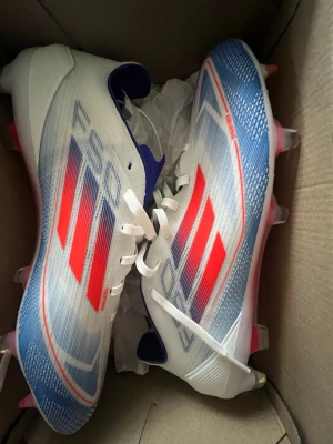 Adidas F50 PROS FOTBOLLSSKOR - Snygga Adidas fotbollsskor med vit bas, blå detaljer och röda ränder. Skorna har snörning, låg profil och lätta material för bästa prestanda på lappt gräs. Yttersulan har röda och vita dobbar för bra grepp. Perfekta för dig som vill sticka ut på planen.