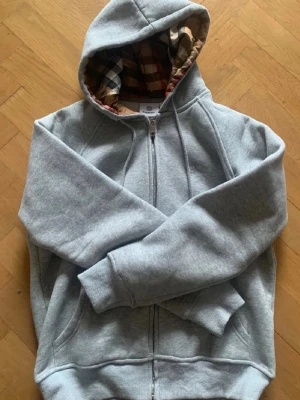 Grå zip-hoodie från Burberry - Snygg grå hoodie från Burberry med dragkedja och klassiskt rutigt foder i huvan. Mjuk bomullskänsla, ribbade muddar och fickor på sidorna. Perfekt för dig som vill ha en stilren och exklusiv look med en touch av brittisk design. Passar S även fast det står M