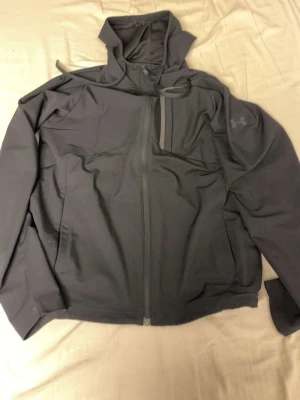 Svart träningsset från Under Armour - Snyggt svart träningsset från Under Armour med jacka och byxor i mjukt funktionsmaterial. Jackan har hög krage, hel dragkedja och bröstficka med dragkedja. Byxorna har dragsko i midjan och diskret logga på låret. Perfekt för träning eller chill.