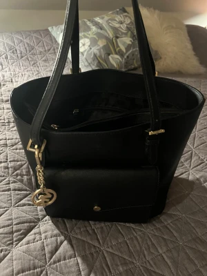 Svart handväska från Michael Kors - Snygg svart handväska från Michael Kors i slätt läder med guldfärgade detaljer och en dekorativ MK-berlock. Väskan har dubbla axelremmar, en framficka med knapp och dragkedja upptill. Perfekt för dig som vill ha en stilren och rymlig väska.