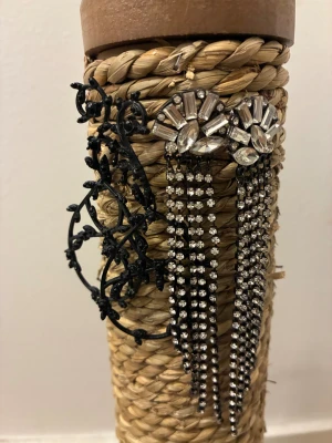 Två par statement örhängen - Säljer två par coola örhängen. Ett par är långa, glittriga och silverfärgade med strass och hängande kedjor, perfekta för att sticka ut. Det andra paret är svarta, stora och har en snygg blommig design med detaljerade blad. Paketpris 199kr styckpris 129kr. 