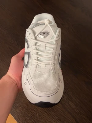 Vita Dior sneakers med reflekterande detaljer - Säljer ett par vita Dior sneakers med reflekterande detaljer på sidan och svart logga. Skorna har en chunky sula, meshpaneler för extra ventilation och vita skosnören. Snygg design med futuristiska vibbar och tydlig Dior-märkning på plösen.