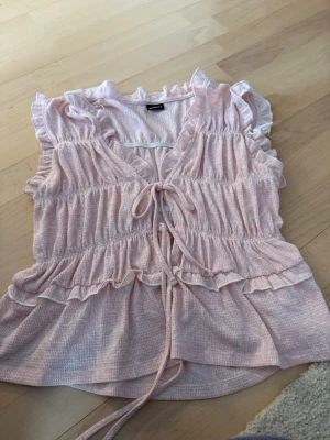 Ljusrosa volangtopp från Gina Tricot - Supersöt ljusrosa topp från Gina Tricot med volanger och knyt framtill. Toppen har v-ringning, korta ärmar med volangkant och ett lätt, luftigt tyg som passar perfekt till varma dagar.