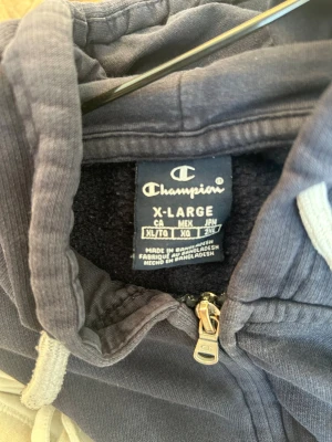 Mörkblå hoodie från Champion XL - Mörkblå hoodie från Champion i storlek XL. Klassisk modell med dragkedja framtill, justerbar huva med vita snören och mjukt material på insidan. Perfekt för en avslappnad och sportig stil. Metallzip med Champion-logga.