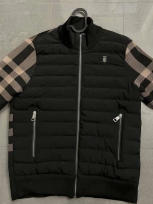 Svart pufferjacka från Burberry - Snygg svart pufferjacka från Burberry med klassiskt rutmönster i beige och svart på ärmar och rygg. Jackan har två dragkedjefickor framtill, hög krage och diskret Burberry-logga på bröstet. Perfekt för dig som vill sticka ut med stilren design och exklusiv känsla.