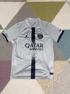 PSG grå matchtröja Jordan x Nike - Snygg grå PSG matchtröja med svart vertikal rand och svarta detaljer. Tröjan har korta ärmar, rund hals och Paris Saint-Germain-logga på bröstet. Tillverkad i lätt och ventilerande funktionsmaterial, perfekt för fotboll eller träning.