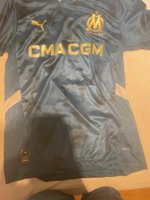 Blå Marseille matchtröja Puma - Snygg blå matchtröja från Olympique de Marseille med guldfärgade detaljer och logga. Tillverkad i lätt och ventilerande material med DryCell-teknologi. Stora tryck på bröstet och klassisk sportig passform.