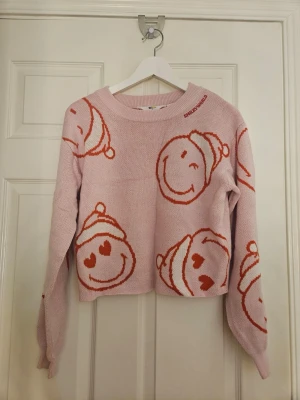 Rosa stickad tröja med smileys - Säljer en ljusrosa stickad tröja från SMILEY WORLD med röda och vita smiley-ansikten i julmössa över hela tröjan. Tröjan har rund hals, långa ärmar och en något croppad passform. Perfekt för dig som gillar lekfulla och färgglada plagg.