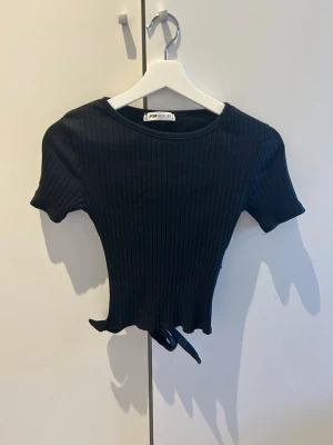 Svart ribbad croppad topp med knyt - Säljer en svart ribbad croppad topp. Toppen har korta ärmar och ett snyggt knytband i ryggen.