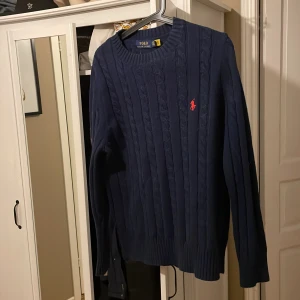 Mörkblå kabelstickad tröja från Polo Ralph Lauren - Säljer en klassisk mörkblå kabelstickad tröja från Polo Ralph Lauren med rund halsringning och röd broderad logga på bröstet. Perfekt för lager-på-lager och stilrena outfits.jag fick den för några veckor sen knappt använt.