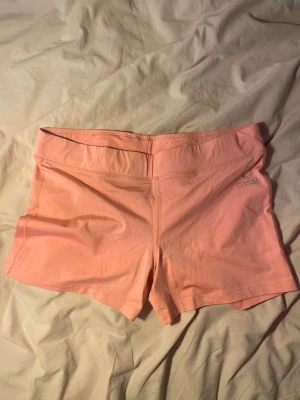 Ljusrosa träningstights från Casall - Säljer ett par ljusrosa träningstights från Casall. De är korta, tighta och har en bred midja för extra komfort. Perfekta för gymmet eller löpning. Materialet känns mjukt och stretchigt, troligen en blandning av bomull och syntet.