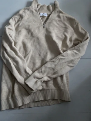 Beige stickad tröja Selected Homme - Säljer en beige stickad tröja från Selected Homme med halv dragkedja och ribbade muddar. Tröjan har en avslappnad passform och är tillverkad i ett mjukt, strukturerat material. Perfekt för lager på lager och enkel att matcha med olika outfits. Använd få gånger och är i fint skick!