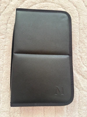 Svart sminkborst väska från morphe - Stilrent svart necessärfodral i konstläder med präglat M på framsidan. Fodralet har dragkedja runtom och flera genomskinliga innerfickor med tryckknappar för smart förvaring av småsaker eller sminkverktyg. 