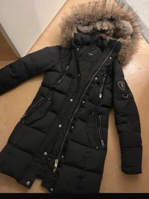 Svart lång dunjacka med fuskpäls - Snygg svart lång dunjacka från Rockandblue med avtagbar huva och fuskpäls. Jackan har flera dragkedjeförsedda fickor, quiltad design och emblem på ärmen. Perfekt för kalla dagar och ger en riktigt cool streetlook. Nypris 5000kr