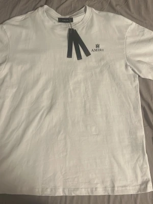 Vit t-shirt från Amiri - Säljer en stilren vit t-shirt från Amiri med svart logga tryckt på bröstet och en diskret detalj på ryggen. Klassisk passform och rund halsringning. Perfekt till jeans eller shorts för en clean look. Passar även M alltså size up är den. Aldrig använd.