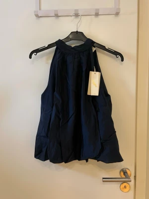 Mörkblå topp med rosett - Snygg mörkblå topp med hög hals och stor rosett man knyter i nacken. Toppen har en luftig, voluminös passform och är ärmlös. Perfekt för dig som vill ha en trendig och elegant look.