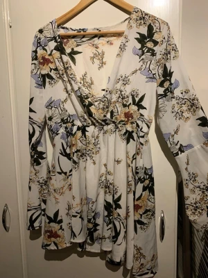 Blommig omlottklänning med v-ringning - Säljer en vit kortklänning med blommigt mönster i gult, lila och grönt. Klänningen har lång ärm, omlott-design och v-ringad hals. Tyget är mjukt och faller fint, perfekt för vår och sommar. Ingen synlig logga på märket.