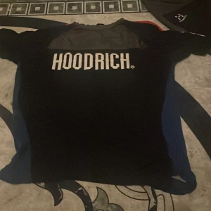 Svart HOODRICH t-shirt med tryck - Svart t-shirt från HOODRICH med stort vitt logotryck på bröstet. Klassisk passform med rund hals och korta ärmar. Perfekt för en streetwear-look och enkel att matcha med jeans eller shorts.