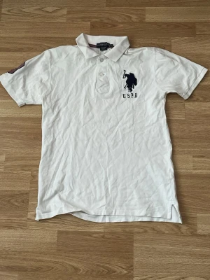 Polo tröja  - Vit pikétröja från USPA med klassisk krage och knappar framtill. Tröjan har korta ärmar och en stor mörkblå logga broderad på bröstet. Perfekt för en avslappnad och sportig stil.